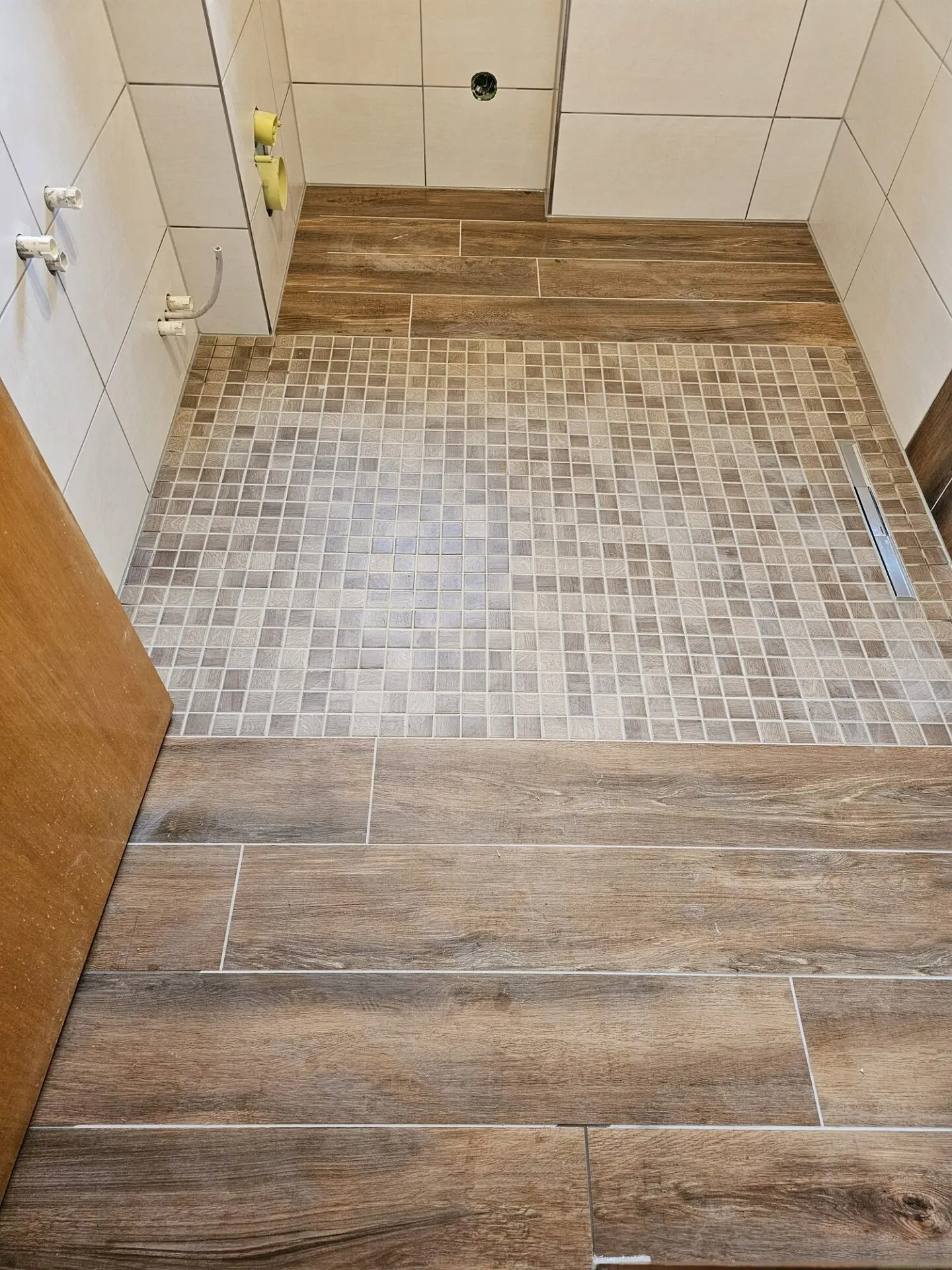 Bodengleiche Dusche mit Mosaikboden und angrenzenden Bodenfliesen in Holzoptik.