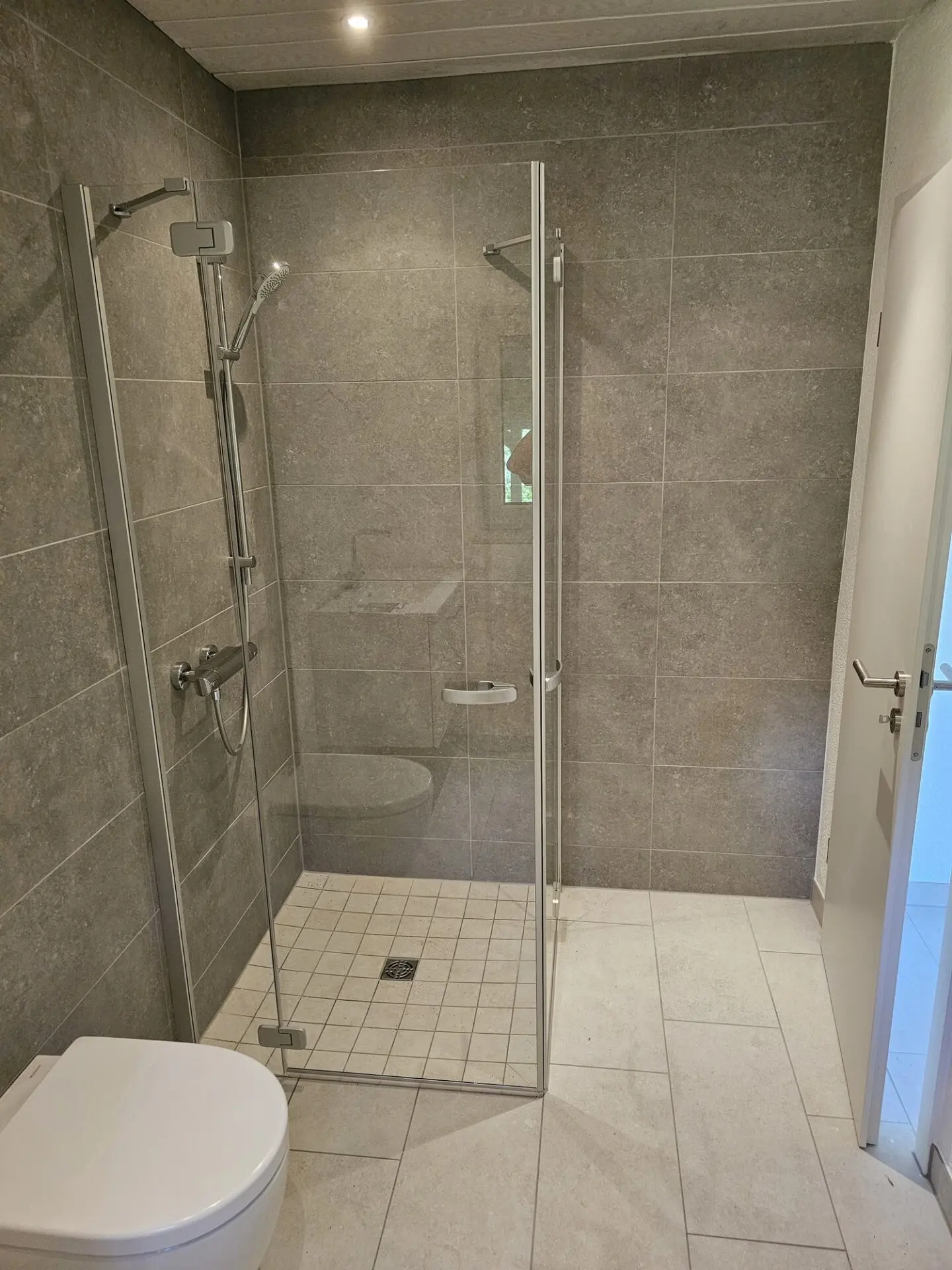 Modernes Badezimmer mit grauen Wandfliesen, hellem Boden und verglaster Dusche.