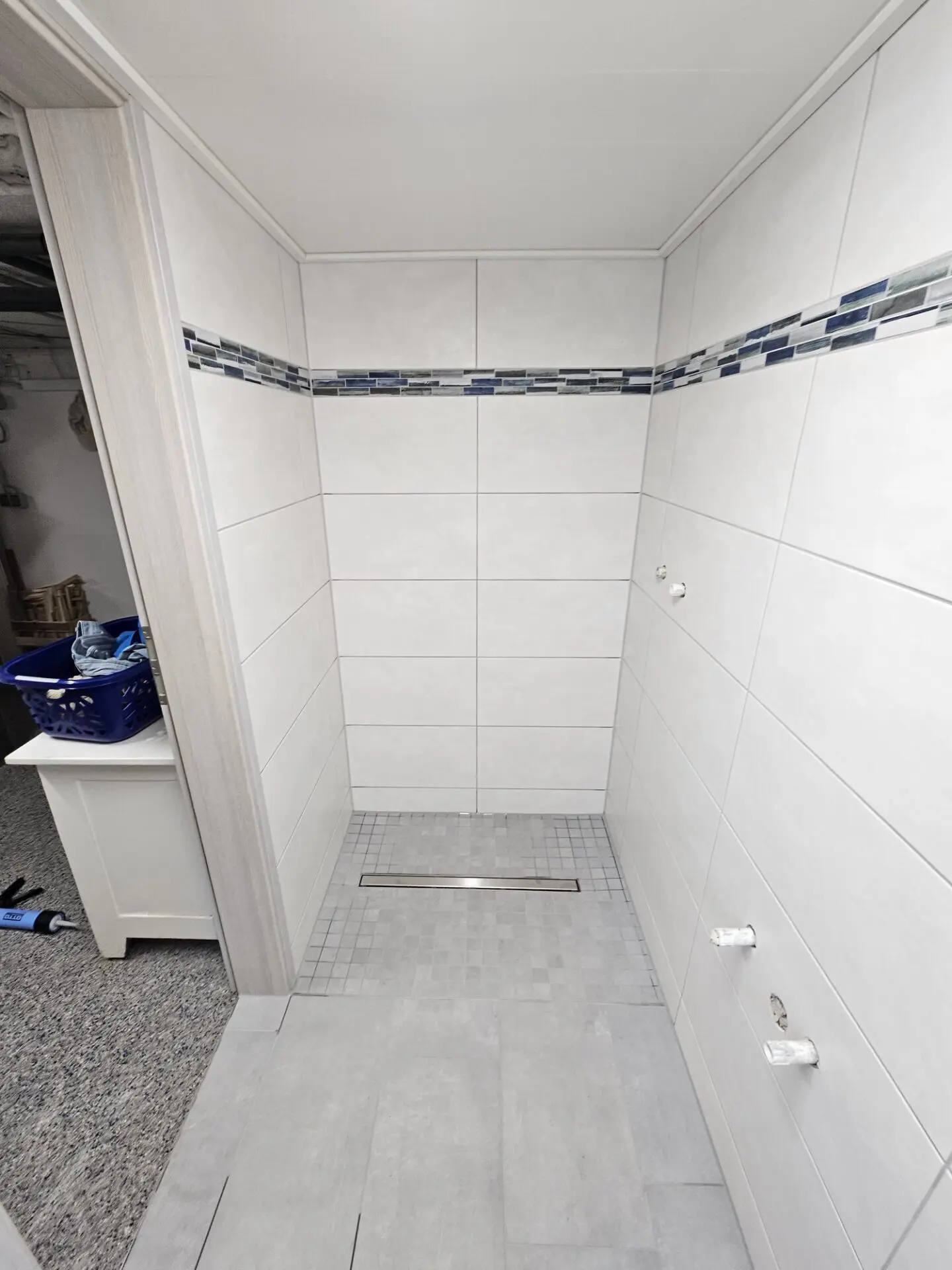 Bodengleiche Dusche mit hellen Wandfliesen, grauem Boden und blauem Mosaikstreifen.