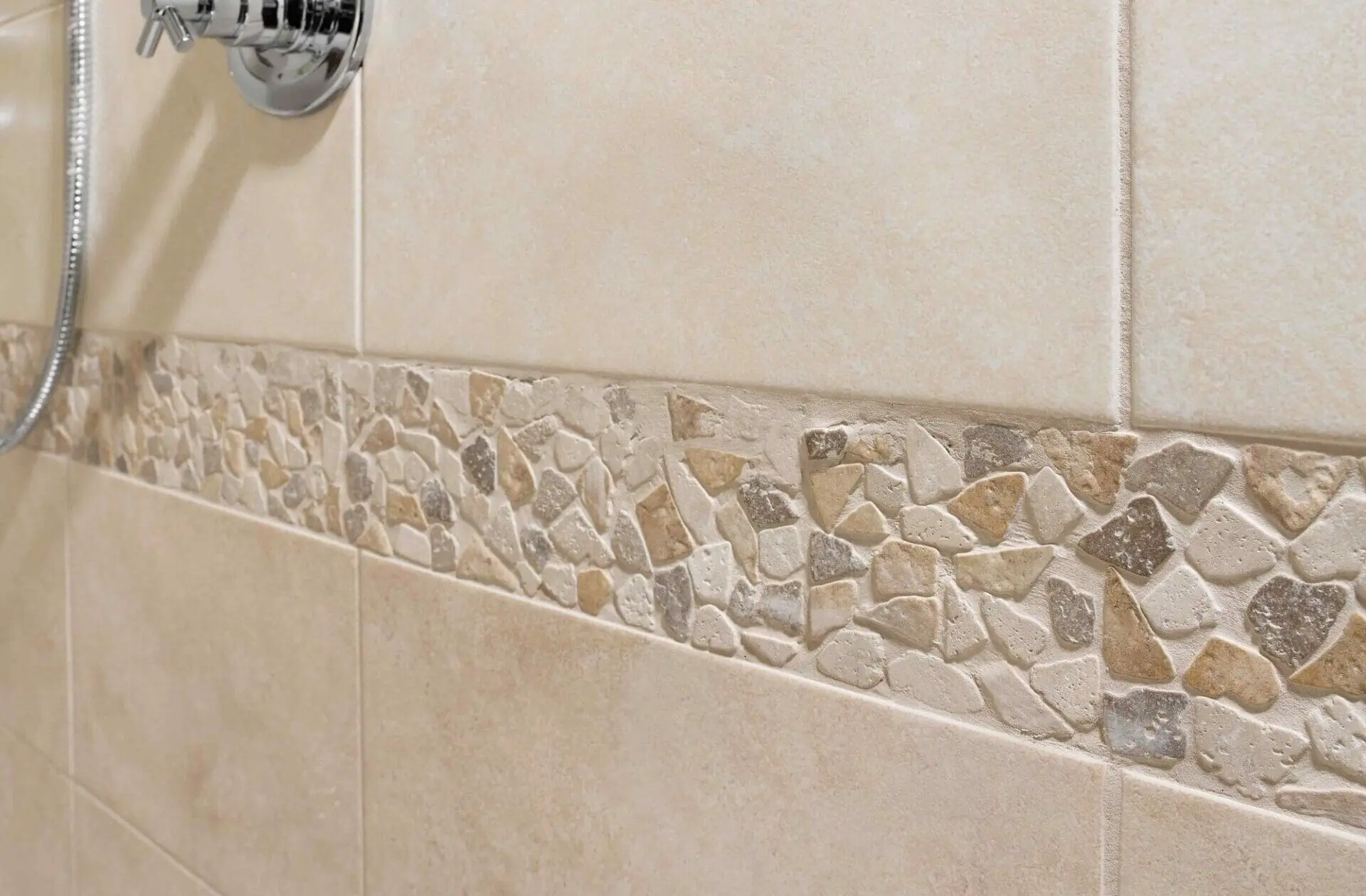 Beige geflieste Duschwand mit Naturstein-Mosaikbordüre und Unterputzarmatur.