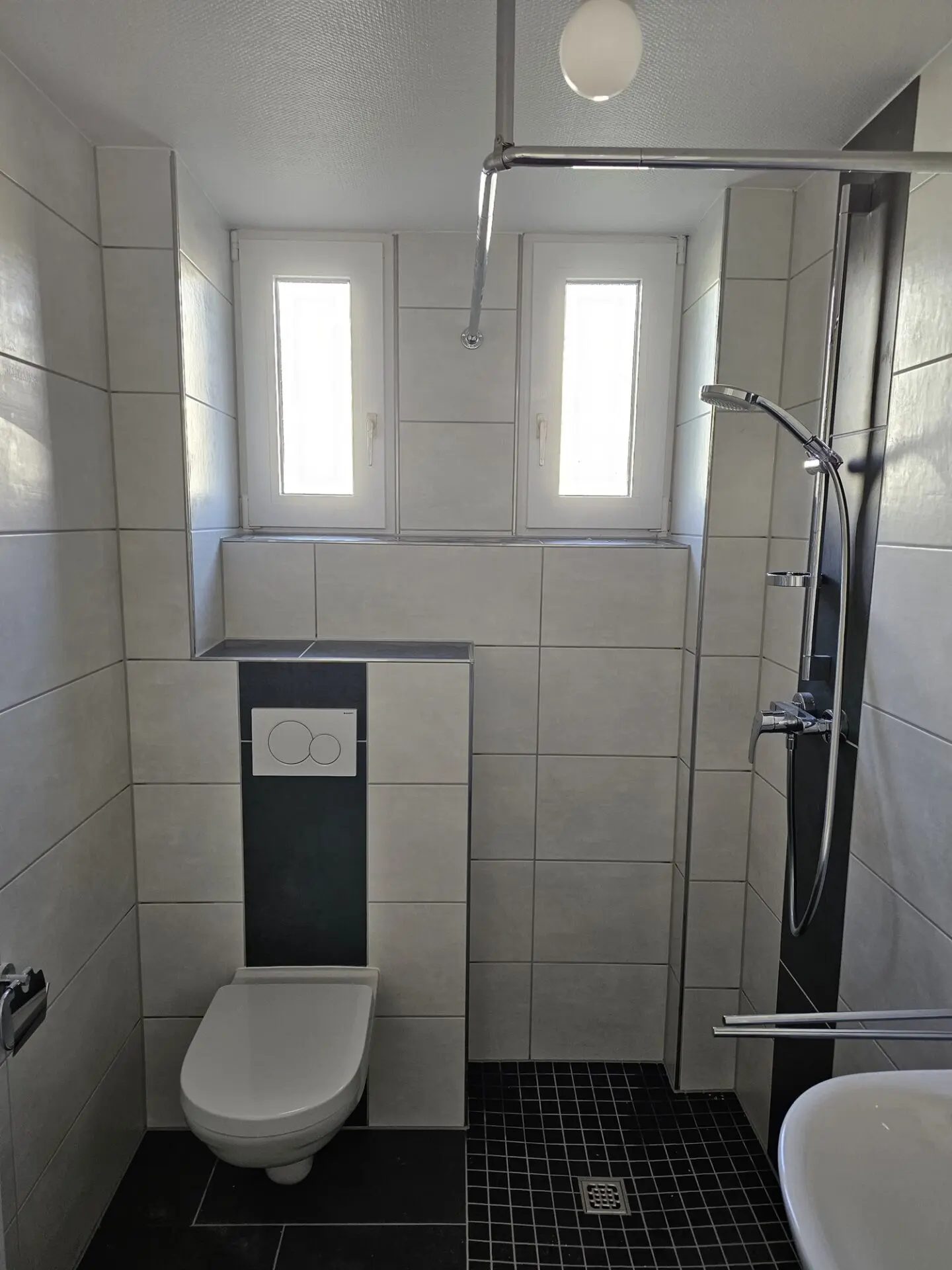 Kleines Badezimmer mit wandhängendem WC, Dusche, grauen Wandfliesen und dunklem Mosaikboden.