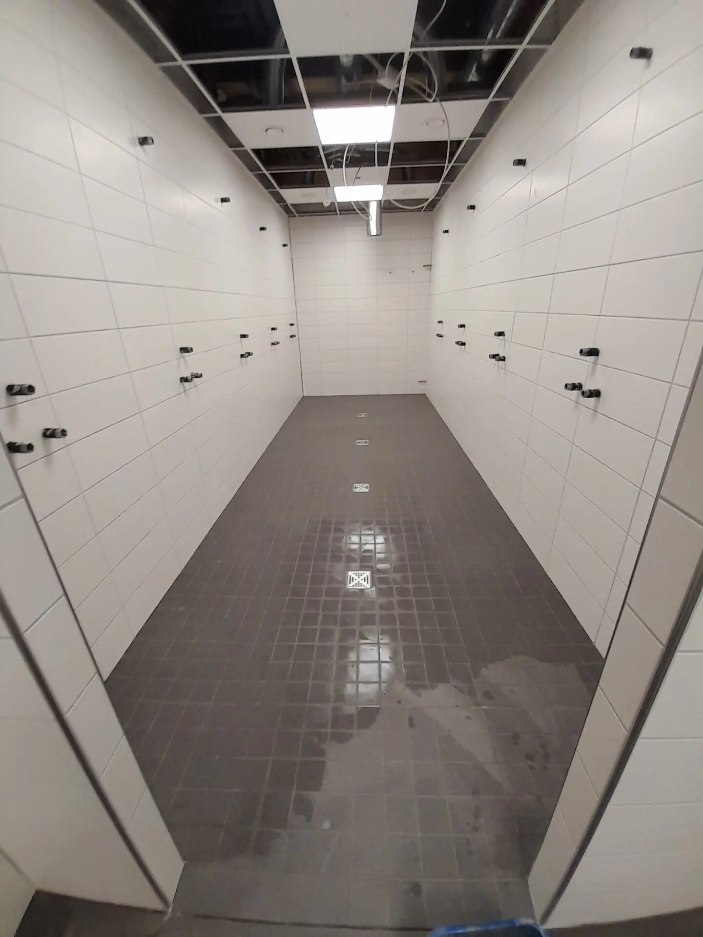 Gefliester Sanitärraum mit weißen Wandfliesen, grauem Mosaikboden und mehreren Wasseranschlüssen.