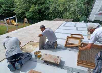 Drei Fliesenleger verlegen großformatige helle Fliesen auf einer Terrasse.